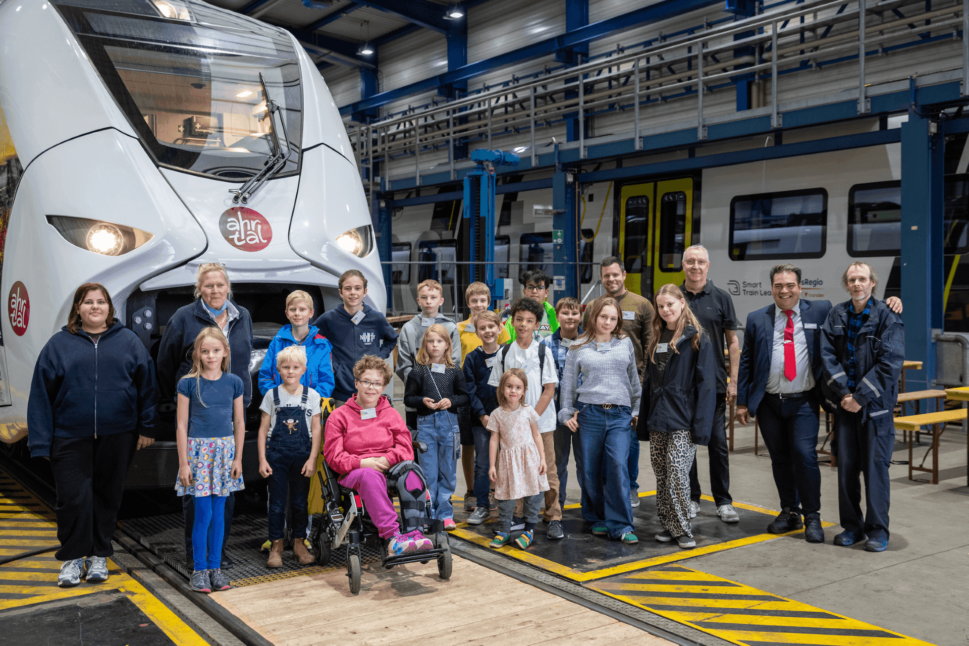 Kinder und Erwachsene stehen vor einem weißen Regionalzug in einer Werkhalle. In der Gruppe befindet sich auch eine Person im Rollstuhl. Im Hintergrund sind weitere Schienenfahrzeuge und technische Anlagen der Zugwartung zu sehen.